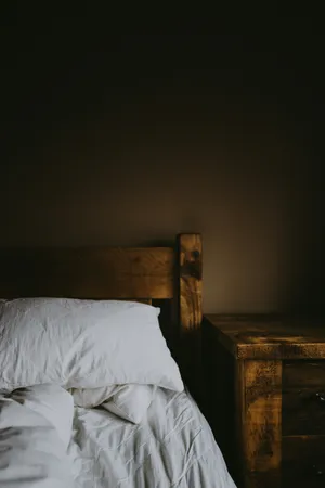 Wooden bed with headboard, white pillow, and wrinkled white duvet next to a wooden nightstand against a dark wall.