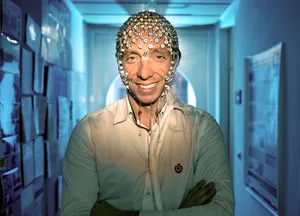 Wissenschaftler mit verschränkten Armen, weißem Hemd mit kleinem Emblem, steht in einem Flur mit blauem Licht und an den Wänden hängenden Plakaten.