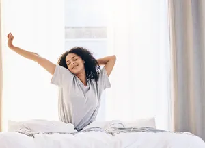 Person mit lockigem Haar in grauem T-Shirt sitzt auf einem Bett und streckt die Arme nach oben.
