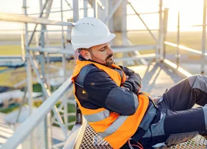Person mit weißem Schutzhelm und orangefarbener Warnweste lehnt mit verschränkten Armen auf einem Metallgeländer.