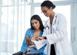 Ärztin in weißem Kittel mit Stethoskop zeigt sitzender Patientin in blauer Bluse und grauem Top Informationen auf Smartphone.